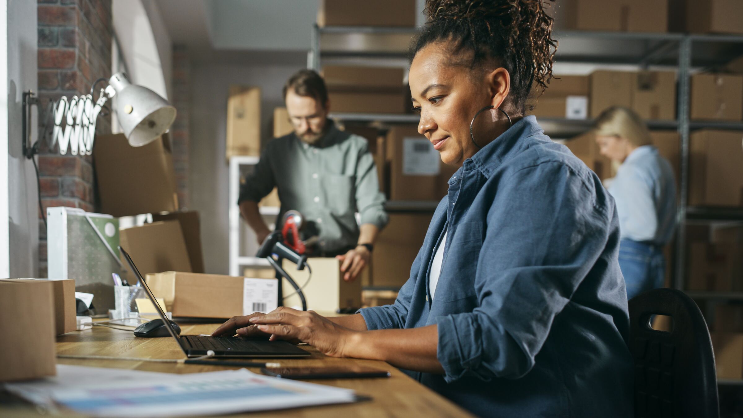 A small business owner working at a laptop in her shop, with a colleague packing orders behind her